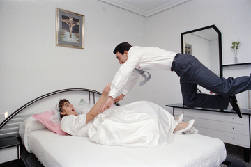 Zu sehen ist ein Ehepaar in einem Schlafzimmer. Die Braut liegt auf dem Bett und der Mann springt zu ihr. Beide freuen sich. © Juan de la Cruz Megias Mondéjar
